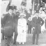 Image: woman in white dress and hat standing among three men in tophats and tails