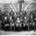 Image: A group of men attired in nineteenth-century South Australian customs service uniforms pose for a photograph. A large Union Jack flag appears as a backdrop behind the men