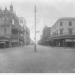 Image: a wide intersection with a lamppost in the centre. On one corner is an ornate three storey building with a verandah, balcony and highly decorative and detailed parapet.