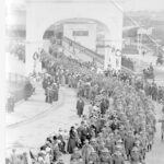 Image: large group of men in uniform walk through archway and along path