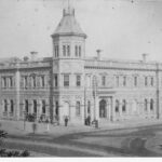 Image: A large, two-storey stone building in Victorian Italianate style. It features a large, octagonal tower with an additional storey in one corner. A group of men in nineteenth century attire congregate in front of the building