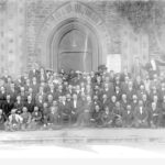 Image: Black and white group photograph of over 150 people. There are five rows of people, mostly men, standing in front of a church