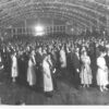 Image: a crowd of men and women wearing 1920s era clothing stand in pairs in a large hall the ceiling of which is decorated with lines of small lights.