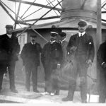 Image: A group of middle-aged Caucasian men in early Edwardian attire pose for a photograph outside a metal lighthouse. One man is wearing what appears to be a government uniform