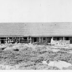 Image: A group of Caucasian workmen in early Edwardian attire sit on scaffolding in front of a long stone building. The building is divided into three separate dwellings with verandahs