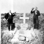 Image: Two middle-aged Caucasian men in Edwardian attire flank a simple grave site. The grave is covered with stone and features a large white wooden cross, to which is attached a sign that reads ‘Sacred to the memory of G. Marment, drowned July 28 1910’