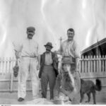 Image: A group of young Caucasian men and one young Caucasian woman in early Edwardian attire pose for a photograph in front of a cottage and white picket fence