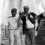 Image: Two young moustachioed Caucasian men in Edwardian-era work clothing pose for a photograph. The men stand in front of a cottage and white picket fence, and the man on the left holds a large crayfish in his left hand