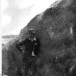 Image: A young moustachioed Caucasian man in casual early Edwardian attire poses for a photograph next to a large rock formation on an island