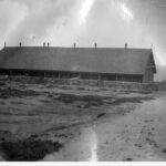 Images: A long stone building surrounded by a low stone fence sits on raised ground overlooking the sea. Part of a coastal land-form is visible in the distance behind the building