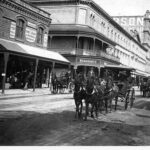 Image: horse drawn vehicles travel down a dirt road lined with terraced shops