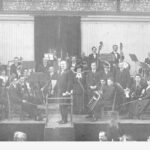 Image: A large symphonic orchestra is assembled on a stage at a large exhibition hall