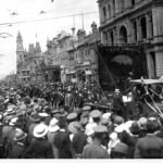 Image: crowd gathered to watch a procession along a street, large banner in centre