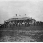 Image: Black and white photograph of a gathering of people. Some are standing on the verandah of a house. Another groups stands further back from the house (closer the camera) in a large circle, joined by their hands