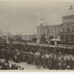 Image: A large group of people in 1880s dress stand in distinct lines in front of a large building with a domed roof which is decorated with flags and bunting.