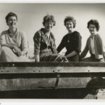 Image: Four smiling women seated on a metal object