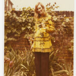 Image: A woman posing for a photograph outside. Behind her is a brick wall covered in vines. She is holding a yellow flower and smiling at the camera.