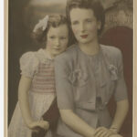 Image: A colour photograph of a young child and her mother. The mother is sitting on a chair, the girl is standing to her left