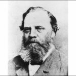 Image: A photographic head-and-shoulders portrait of a middle-aged Caucasian man with a bushy salt-and-pepper beard and receding ear-length hair. He is wearing a late-Victorian suit with bowtie