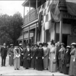 Image: A group of women and men in Edwardian attire stand in front of a two-storey stone building with flags hanging from the second-floor verandah