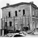 Image: A two-storey stone building with burned out windows and melted roof cladding. The burnt remains of another building are in the foreground