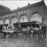 Image: A horse-drawn cart passes in front of a large brick building with the words ‘Federal Hall’ in large letters on its front facade