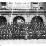 Image: A large group of people sit in rows outside the grand arched entrance of a large building, posing for a formal photograph. Three more people lean more casually on the balustrade of a balcony above.