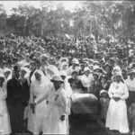 Image: Black and white photograph of a large crowd standing behind a group of women dressed in white and a man in a dark suit. In the centre of the picture a wounded soldier can be seen in a stretcher bed.