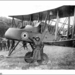 Image: A man in a First World War-era flying outfit stands in front of a British Royal Air Force biplane