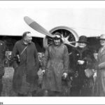 Image: A man in First World War-era aviator attire stands in front of an aircraft with two men and two women