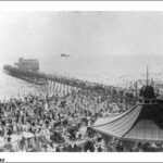 Image: A large crowd of people watch an early twentieth century aircraft make a low pass over a jetty near a seaside community