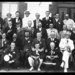 Image: A group of Caucasian men of varying ages dressed in Edwardian-era suits or uniforms pose for a photograph in front of a bluestone building