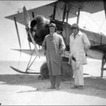 Image: Two men wearing suits, overcoats and early-twentieth century aviator helmets pose for a photograph in front of a biplane