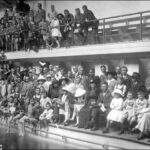 Image: A crowd of people in early twentieth century attire are gathered at the side of a large swimming pool