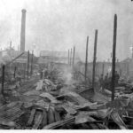 Image: A small group of people stand within the smouldering remains of a building. Charred wooden uprights and collapsed corrugated metal sheeting are all that remains of the structure