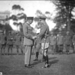 Image: Two men in military uniforms shake hands on an open field. A group of Australian Army soldiers stand in a line in the background