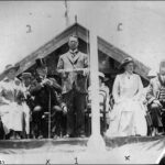 Image: A man in glasses stands at a podium delivering a speech. A group of male and female dignitaries sit on seats on the stage behind the speaker. The roof of a building is visible behind the stage