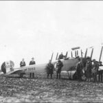 Image: A group of men and one woman stand around a biplane. The identification number ‘H3033’ is painted on both the tail and side of the aircraft