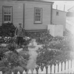 Image: A man in uniform stands in a flower garden next to a cottage