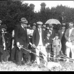 Image: A man in a hat and suit operates a plow in front of a small group of people