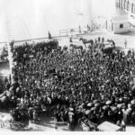 Image: A large crowd of men gather on a wharf to hear a speech