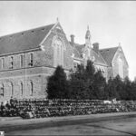 Image: Hundreds of schools students in black uniforms pose in lines outside a large symmetrical stone building with decorative brickwork between floors, two huge arched windows on the front gable ends and smaller windows on the sides.