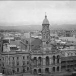 Image: A large stone building with a central tower capped by a dome. Cars drive along a street in front of the building