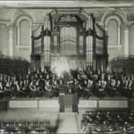 Image: An orchestra poses for a photograph on a stage in front of a large pipe organ