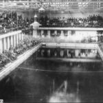 Image: A crowd of people in early twentieth century attire are gathered at the side of a large swimming pool