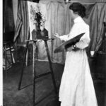 Image: A young Caucasian woman in a full-length Edwardian-era dress paints a still life on an easel. The painting features a couple of vases holding flowers