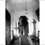 Image: the entry hall of a house. The decorative tiled floors are covered by a runner carpet which extends through an archway supported by columns and up a narrow staircase.