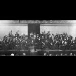 Image: A large symphonic orchestra is assembled on a stage at a large exhibition hall