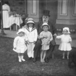 Image: Black and white photograph of seated man, wearing a top hat, surrounded by four small children. In the background a woman stands near a table facing away from the camera.