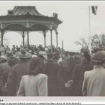 Image: a crowd of people, some in military uniform, watch dignitaries give speeches from a rotunda. A flag, possibly the Union Jack, is just visible on a flagpole to the right of the photograph.